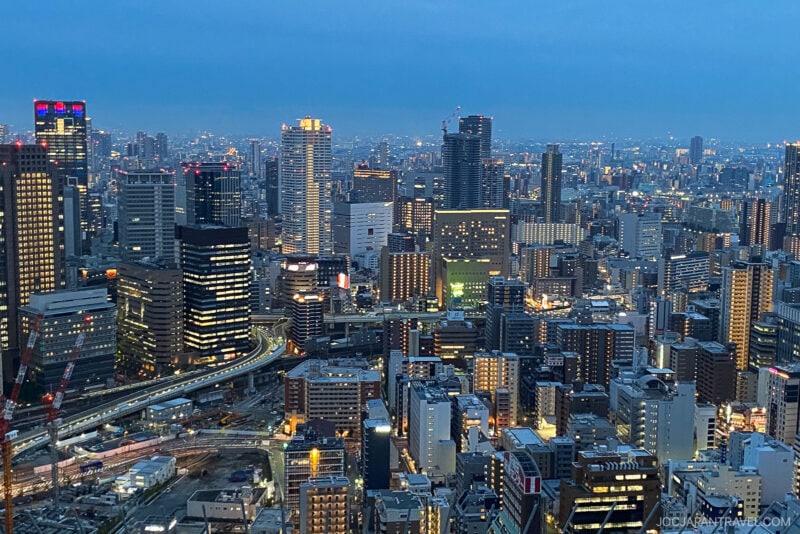 A cityscape at dusk with numerous high-rise buildings illuminated, elevated highways, and dense urban structures under a blue evening sky, featuring the striking Umeda Sky Building in the skyline.