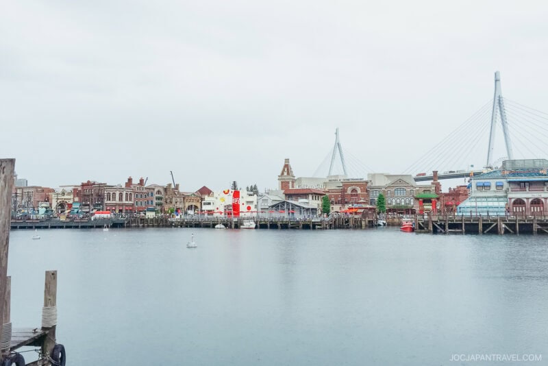 A waterfront view of a city with colorful buildings, a dock, and a modern cable-stayed bridge in the background under a cloudy sky, reminiscent of scenes near Universal Studios Japan. The water is calm with a few small buoys visible.