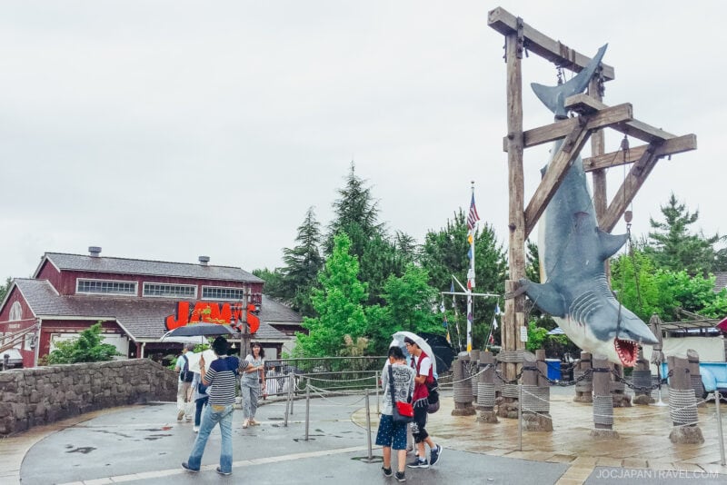 People with umbrellas stand near a large hanging shark statue at the entrance of the Jaws attraction in Universal Studios Japan, with a red-roofed building and green trees in the background on a cloudy day.