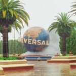 Universal Studios Japan Guide: Everything You Need to Know for Your Visit A large globe with the word UNIVERSAL stands surrounded by palm trees, mist, flowers, and benches at the entrance to Universal Studios Japan on a cloudy day.