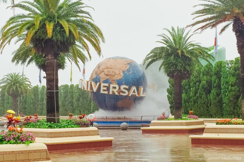 A large globe with the word UNIVERSAL stands surrounded by palm trees, mist, flowers, and benches at the entrance to Universal Studios Japan on a cloudy day.
