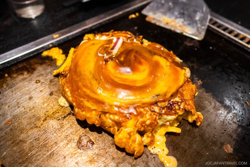 A close-up of a cooked okonomiyaki, a savory Japanese pancake topped with sauce, on a hot griddle in Dotonbori, with a metal spatula nearby.