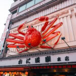 Dotonbori (道頓堀) – Where to Eat in Osaka’s Food Paradise A large red crab sculpture is mounted above the entrance of a seafood restaurant on a bustling Dotonbori street in urban Japan, with buildings and signs visible in the background.