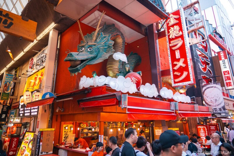 A busy street scene in Japans lively Dotonbori, with people walking past a ramen shop featuring a large green dragon statue above the entrance and bright red signage with Japanese characters.