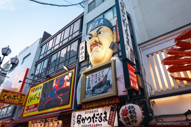 Colorful storefronts in Osaka’s Dotonbori feature a large, dramatic-faced head sign above a restaurant entrance, surrounded by vibrant Japanese signage and billboards on this lively street.
