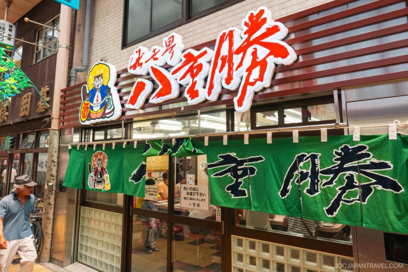 A man walks past Yaekatsu Osaka, a Japanese restaurant with a large red and white sign, cartoon chef illustration, and green fabric banners above the entrance. The scene is bright and lively.
