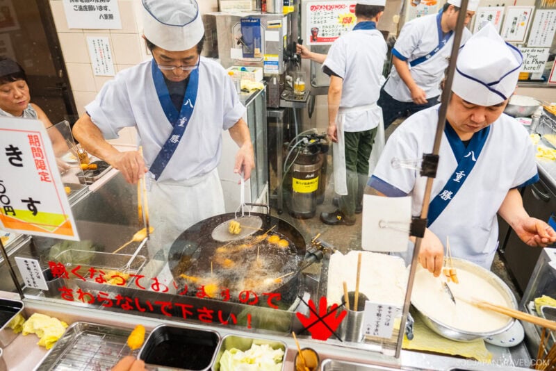 Two chefs in white uniforms and hats fry tempura in the busy Yaekatsu Osaka kitchen. One cooks with chopsticks over hot oil, while the other prepares food at a counter, surrounded by bright signs and kitchen tools.