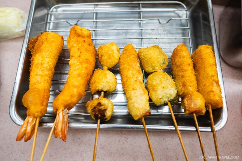 A metal tray at Yaekatsu Osaka holds several skewers of assorted golden-brown deep-fried foods, including shrimp and round vegetables, all coated in crispy breadcrumbs and placed neatly on a wire rack.