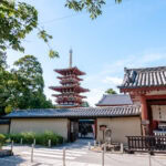 Yakushi-ji (薬師寺): Nara’s Temple of Healing A traditional Japanese temple entrance with a large wooden gate and a tall, red five-story pagoda in the background at Nara Yakushi-ji, surrounded by greenery and under a blue sky.