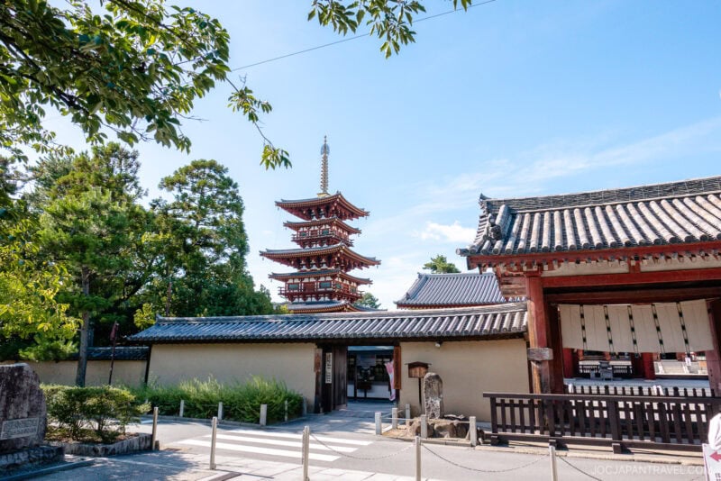 A traditional Japanese temple entrance with a large wooden gate and a tall, red five-story pagoda in the background at Nara Yakushi-ji, surrounded by greenery and under a blue sky.