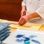 Yamanaka Sushi (やま中) – Omakase Dining Inside Hakata Station A chef in a white coat skillfully slices raw fish on a yellow cutting board at Yamanaka Sushi Fukuoka, with neatly stacked plates and a patterned dish visible in the foreground.