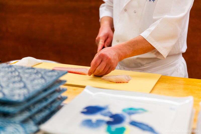 A chef in a white coat skillfully slices raw fish on a yellow cutting board at Yamanaka Sushi Fukuoka, with neatly stacked plates and a patterned dish visible in the foreground.