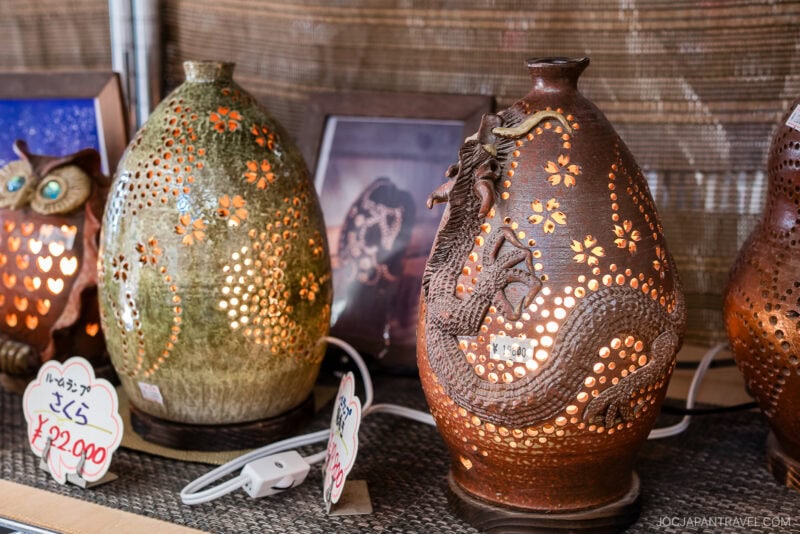 Decorative ceramic lamps from Yomitan Pottery Village, with intricate cut-out patterns, sit on a shelf; one features a dragon design. Warm light glows through the holes, illuminating the details. Price tags and electrical cords are visible nearby.