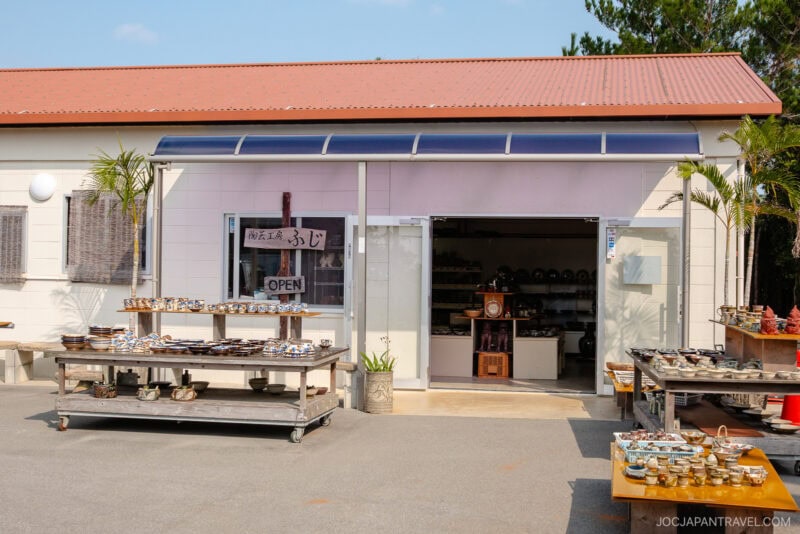 A small pottery shop in Yomitan Pottery Village with tables displaying various ceramic items outside, under a sunny sky. The shop has a red roof, white walls, and an open entrance revealing more pottery inside.
