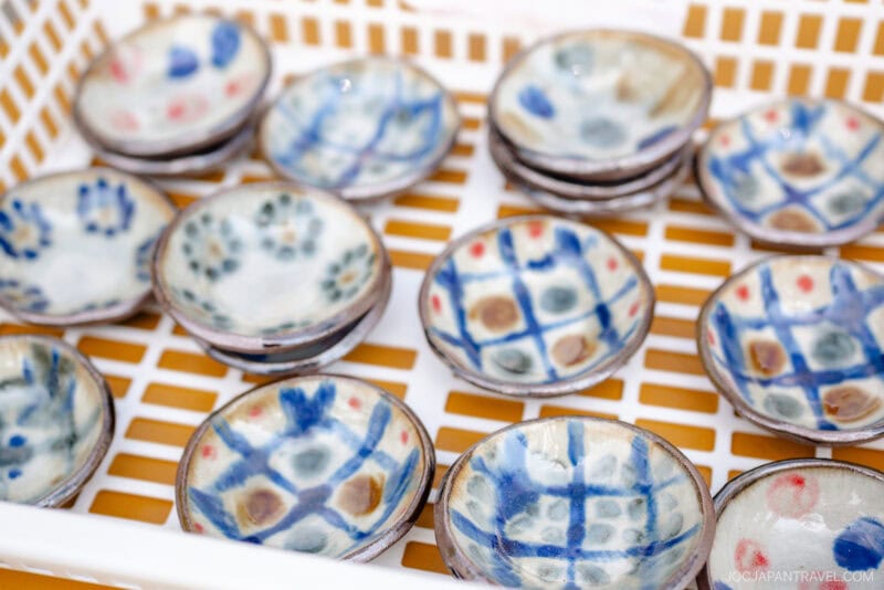 Small ceramic bowls from Yomitan Pottery Village, adorned with colorful geometric and floral motifs in blue, red, and yellow, are neatly stacked in a white plastic basket.