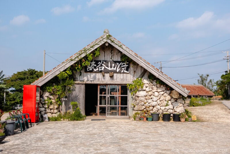 A rustic stone building with a wooden gabled roof stands in Yomitan Pottery Village, green plants hanging around the entrance and Japanese characters on a sign above the door, all under a blue sky with scattered clouds.