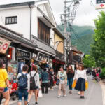 Yufuin Yunotsubo Kaido (湯の坪街道) – A Street Full of Sweets, Souvenirs, and Scenery People walk along Yufuin Yunotsubo Kaido, a lively shopping street lined with traditional buildings, shops, and signs in Japanese. Trees and distant green hills are visible in the background, adding to the area’s vibrant atmosphere.