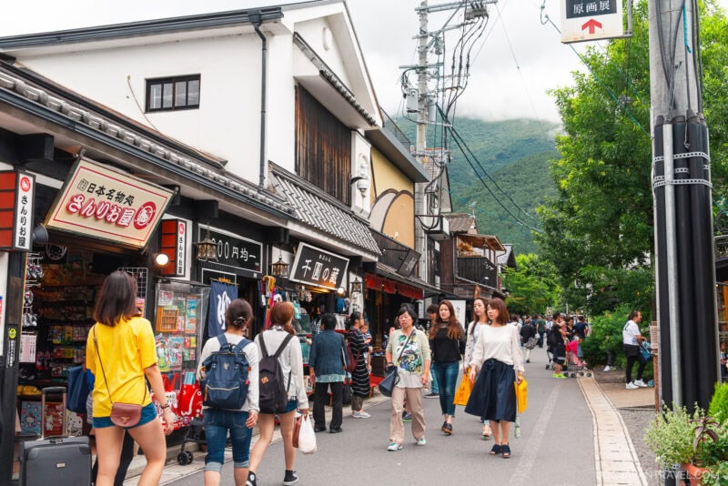 People walk along Yufuin Yunotsubo Kaido, a lively shopping street lined with traditional buildings, shops, and signs in Japanese. Trees and distant green hills are visible in the background, adding to the area&rsquo;s vibrant atmosphere.
