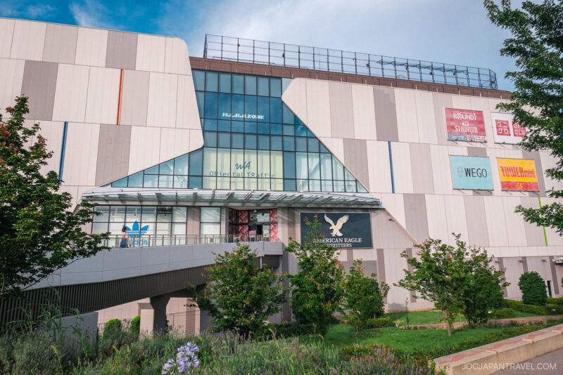 Modern shopping mall exterior with large glass windows, multiple brand signs including Adidas and American Eagle, and a pedestrian bridge surrounded by greenery near Zepp DiverCity Odaiba.