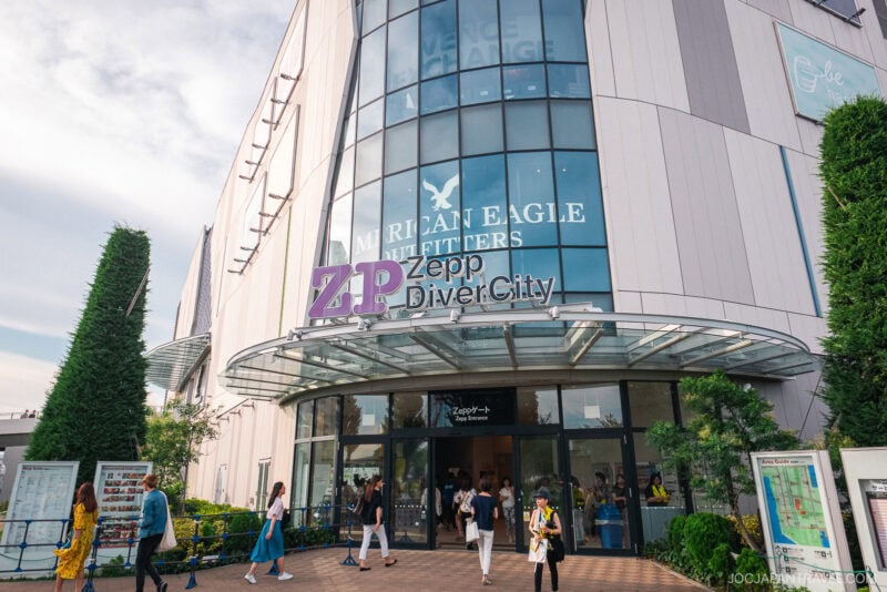 People walk past the entrance of Zepp DiverCity Odaiba, a modern multi-story building with large glass windows, American Eagle signage above, and greenery surrounding the entrance.