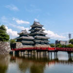 Matsumoto Castle (松本城) – Nagano’s Striking Black Castle Matsumoto Castle, a traditional Japanese landmark with striking black walls, stands beside a moat and is connected by a vibrant red bridge. Trees and modern buildings rise in the background beneath a blue sky dotted with clouds.