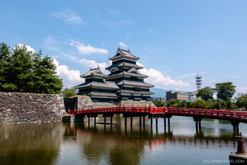 Matsumoto Castle, a traditional Japanese landmark with striking black walls, stands beside a moat and is connected by a vibrant red bridge. Trees and modern buildings rise in the background beneath a blue sky dotted with clouds.