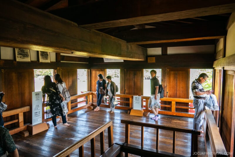 A group of people, some wearing masks, stand and walk inside a traditional wooden Japanese building at Matsumoto Castle, looking out the windows and reading information signs as sunlight filters through and illuminates the wooden interior.