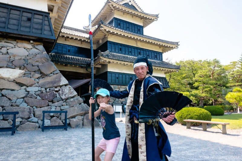 A child poses playfully with a staff next to a costumed man holding a black fan in front of Matsumoto Castle on a sunny day.