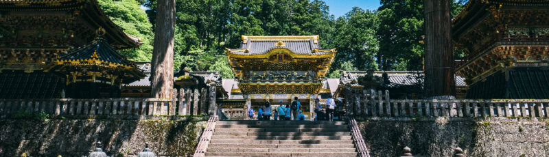 A group of people stands at the base of stone steps leading to an ornate, gold-accented traditional Japanese temple surrounded by tall trees and historic wooden structures.