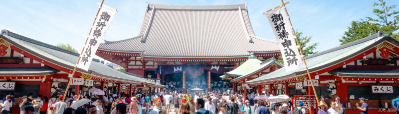A large crowd gathers at a traditional Japanese temple with red buildings, white banners with Japanese writing, and a main hall under a blue sky.