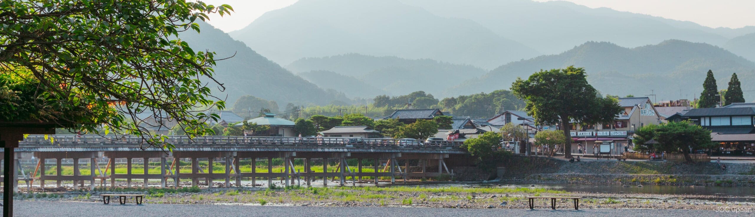 A wooden bridge spans a river in a small town, with traditional houses and lush green trees. Misty mountains rise in the background, creating a peaceful, scenic landscape.