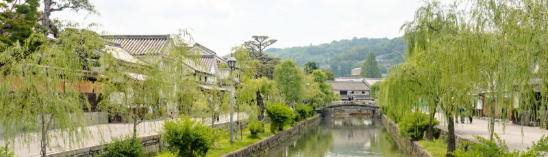 A peaceful canal lined with willow trees, traditional Japanese buildings, and a stone bridge, set against a backdrop of green hills on a bright, clear day.