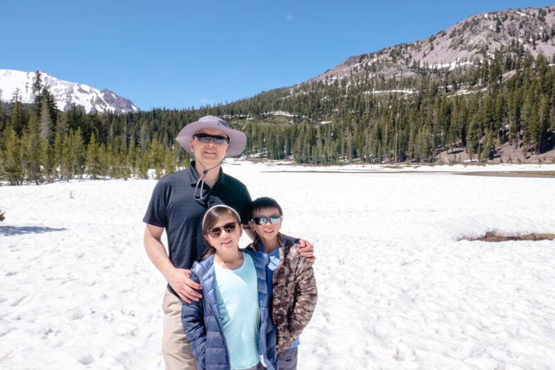 A man and two children stand together on a snowy landscape in Lassen Volcanic National Park, surrounded by pine trees and mountains under a clear blue sky. They are smiling, wearing sunglasses and light jackets—a perfect moment for any travel guide.