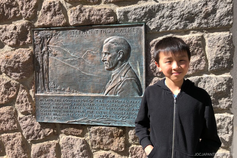 A young boy in a black hoodie stands next to a stone plaque honoring Stephen Tyng Mather, with his profile and mountains engraved, at Lassen Volcanic National Park, affixed to a sunlit stone wall—perfect for any travel guide.