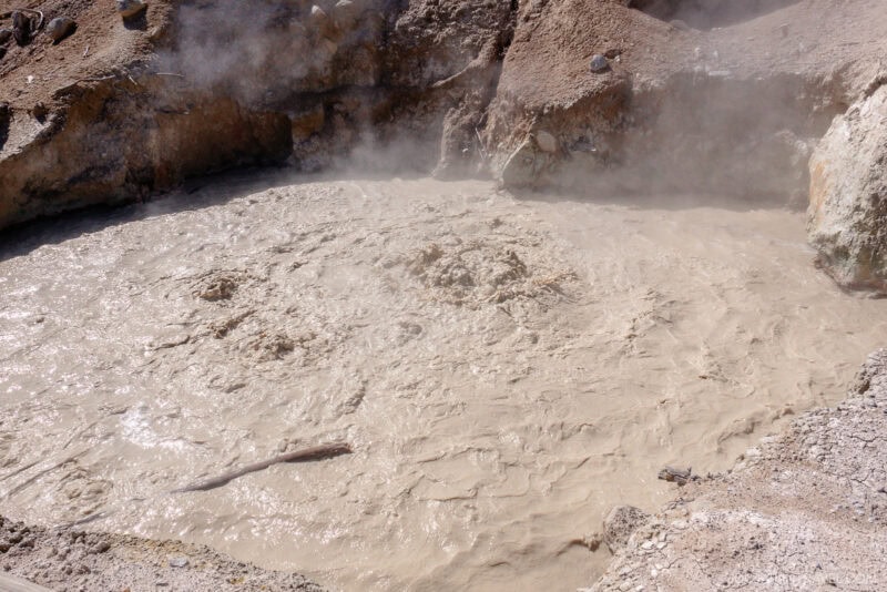 A gray, bubbling mud pool with steam rising from the surface, surrounded by rocky, barren terrain—an otherworldly sight featured in any travel guide to Lassen Volcanic National Park.