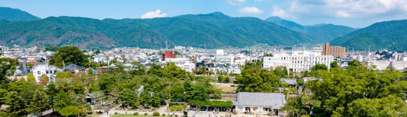 A scenic view of a small city with many trees in the foreground, modern buildings, and green mountains in the background under a blue sky with scattered clouds.