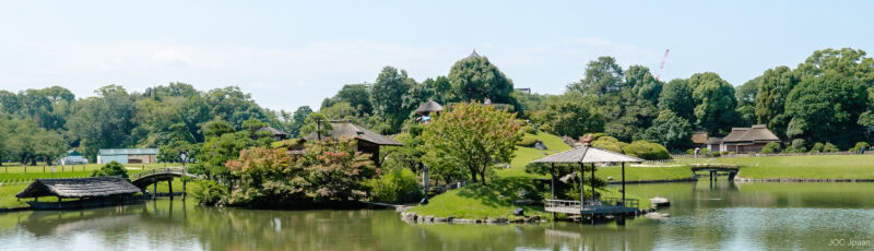 A serene Japanese garden with a pond, wooden bridges, traditional gazebos, and lush green trees under a clear blue sky.