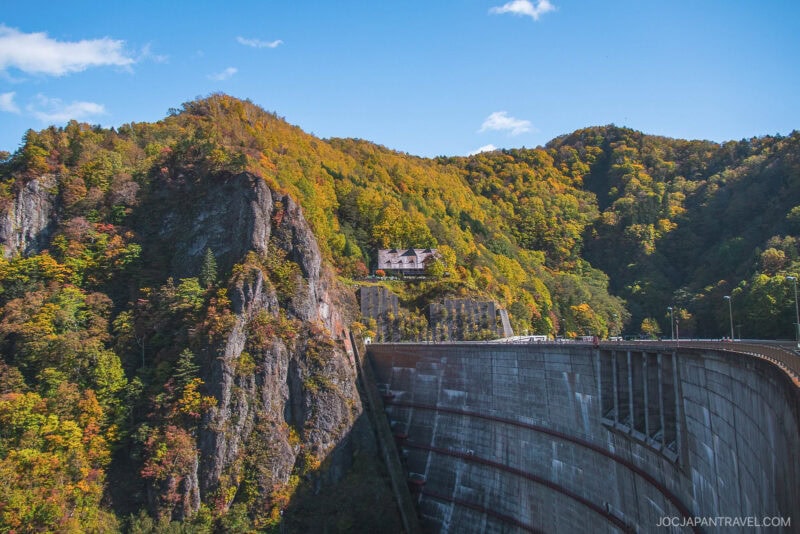 A house sits atop a rocky, tree-covered hill with vibrant Sapporo autumn foliage, overlooking a large concrete dam under a clear blue sky.