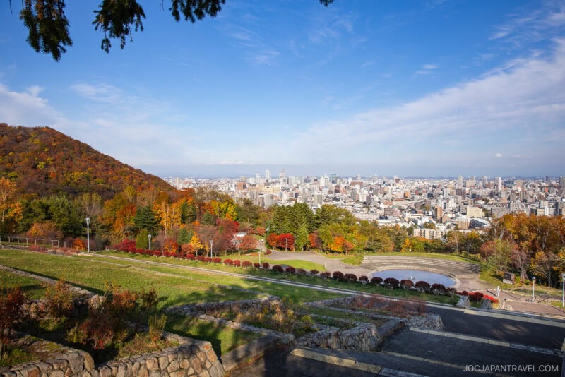 A hillside park with green and sapporo autumn foliage overlooks a cityscape under a blue sky with scattered clouds. Stone steps and pathways wind down the slope toward the city.