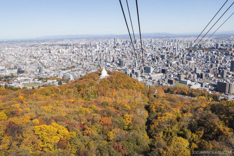 Aerial view of Sapporo city with a cable car line running over a forested hill draped in vibrant Sapporo autumn foliage, transitioning toward urban high-rise buildings in the background under a clear sky.
