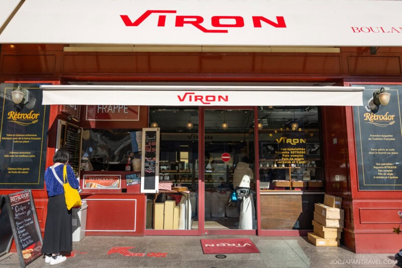 A person with a yellow bag stands in front of Viron, one of the renowned bakeries in Tokyo, known for its French-style bread and pastries, featuring a red storefront, glass doors, and various signs and menus displayed.