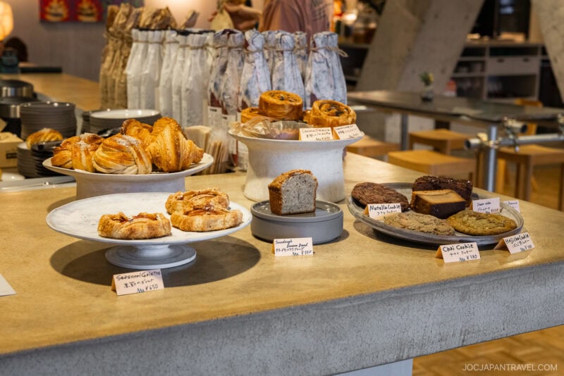 A bakery counter displays an array of pastries, breads, and cookies on plates and stands, each with handwritten labels—a scene reminiscent of the inviting bakeries in Tokyo. The background features a modern, cozy cafe interior.