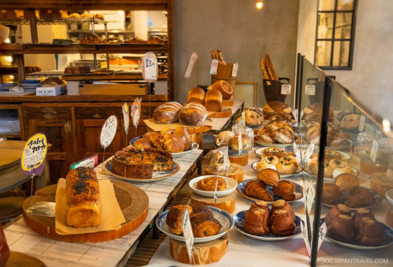 A bakery display inspired by Tokyo bakeries showcases various bread, pastries, and cakes on wooden and ceramic trays behind a glass barrier, with handwritten labels and a cozy interior in the background.