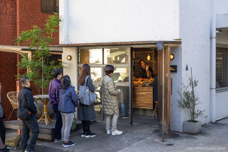 People stand in line outside a small bakery with a wooden door, while a staff member inside waves and smiles; fresh pastries and bread are displayed on the counter near the entrance. This spot is among the beloved bakeries in Tokyo.