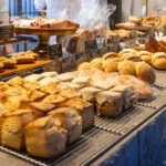 Must-Visit Bakeries In Tokyo A bakery display filled with various types of bread and pastries, including loaves, rolls, and sweet buns, arranged neatly on metal racks and wooden shelves—just what you’d expect from must-visit bakeries in Tokyo, with Japanese labels visible in the background.
