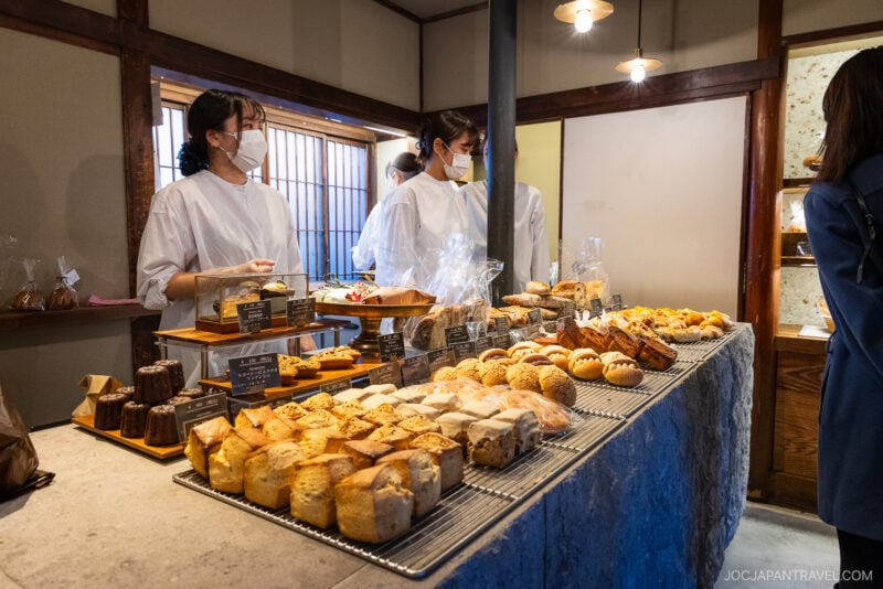 A bakery with two staff in white uniforms and masks stands behind a counter filled with pastries and bread, while a customer looks at the selection in a warmly-lit, rustic interior typical of bakeries in Tokyo.