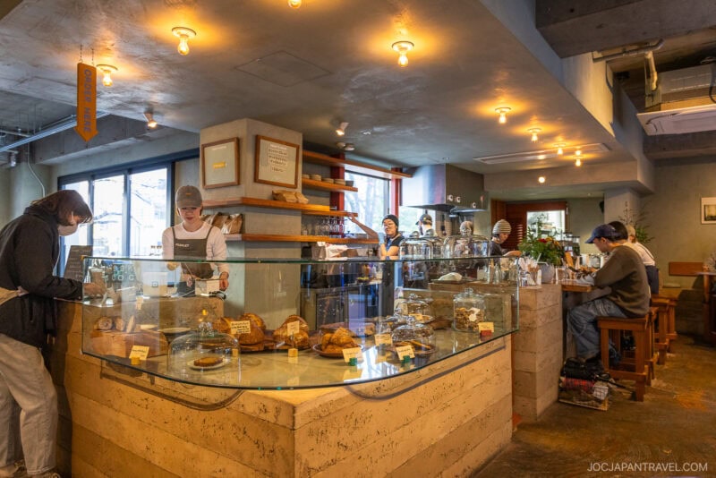 A cozy cafe interior inspired by bakeries in Tokyo; a staff member prepares drinks behind a glass display filled with pastries, while customers order, work on laptops, and chat at tables under warm lighting.