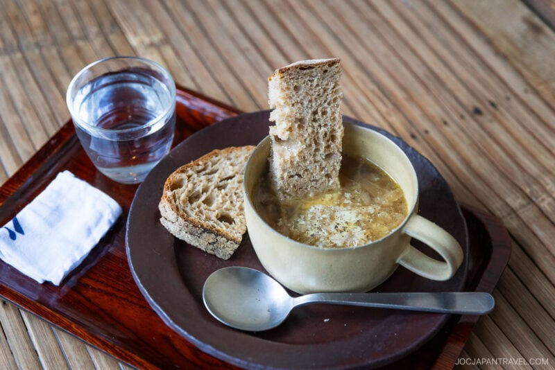 A cup of soup with a slice of rustic bread from bakeries in Tokyo dipped in it, a second slice on the plate, a glass of water, a metal spoon, and a napkin are arranged on a wooden tray atop a wooden table.