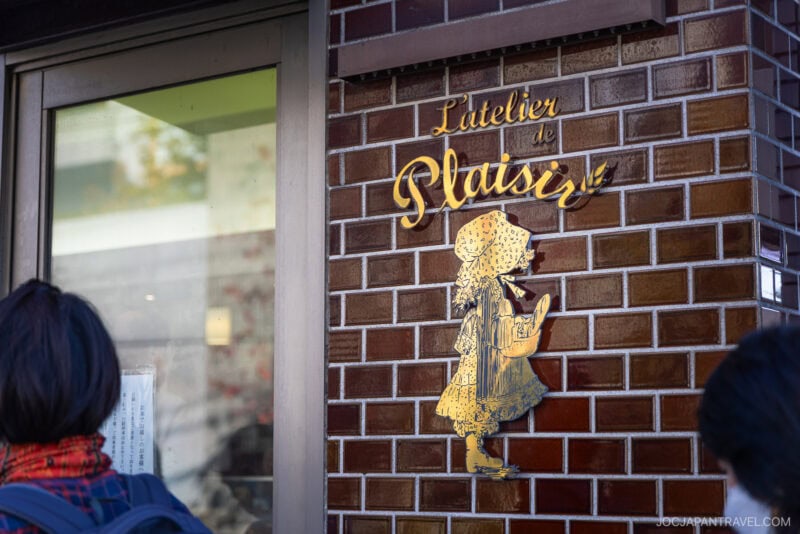 A gold sign reading Latelier de Plaisir with a silhouette of a girl eating bread is mounted on a brick wall next to a door—hinting at the artisanal bread and pastries found in one of Tokyo’s beloved bakeries. Two people are partially visible in the foreground.