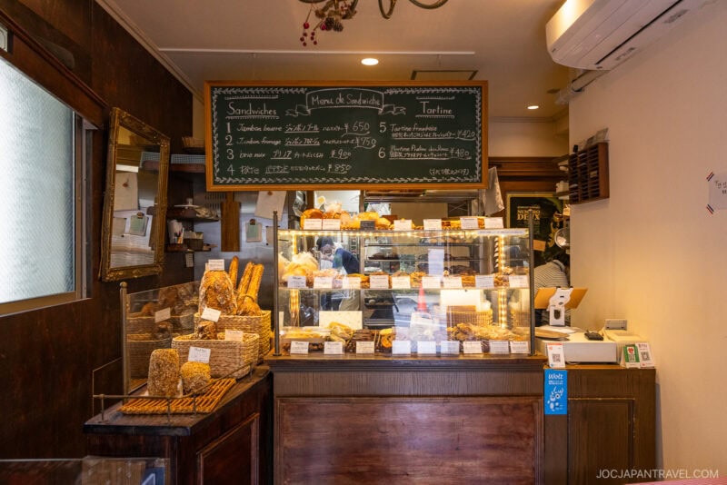 A cozy bakery interior with shelves and a display case filled with fresh bread and pastries. A chalkboard menu hangs above the counter, and sunlight streams in from a window on the left, evoking the charm of bakeries in Tokyo.
